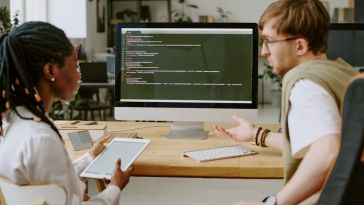 Two software engineers examine code on a computer monitor while working on a project together in an office