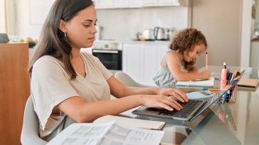 A woman works on a laptop at a dining table while a young child sits in the background drawing in a notebook, illustrating a work-from-home setting.