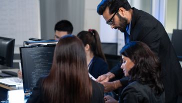 A group of professionals in an office setting work together on a project. A man in a suit leans over, discussing code on a computer screen, fostering collaboration.