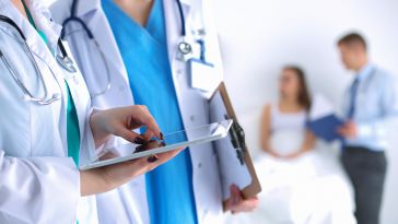 A close up of two doctors wearing white coats, scrubs and stethoscopes while one holds a tablet device.