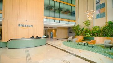 Spacious office lobby with tall wooden walls, glass windows, and vibrant greenery. The "Amazon" logo is visible above a sleek reception desk.