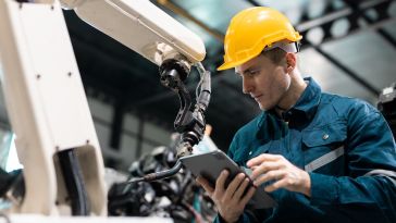 A technician in a yellow hard hat and blue coveralls uses a digital tablet as he works with a large industrial robotic welding arm in a manufacturing facility.