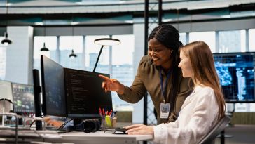 A software engineer points to a line of code on a computer monitor screen while working with another engineer in an office