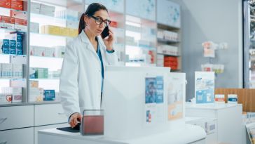 A pharmacist in a white coat talks on the phone behind a pharmacy counter, surrounded by neatly arranged shelves of medicine, conveying a professional setting.