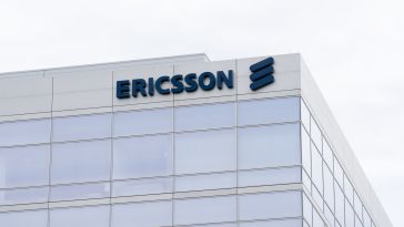 A modern building with a glass facade displays the Ericsson logo in bold blue letters on its corner. The overcast sky adds a neutral backdrop.
