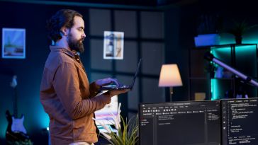 A person stands in front of two monitors with code on them, holding a remote keyboard to show the idea of an engineer working on building an AI tool. 