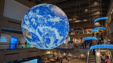 A large, illuminated Earth model hangs in a bustling multi-level science museum. Visitors explore displays, creating a lively and educational atmosphere.
