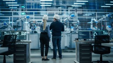 Two engineers in hard hats stand in a modern factory, observing robotic arms in operation. The scene is high-tech, organized, and focused on automation.