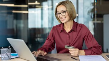 Smiling woman in glasses and red shirt, sitting at office desk with laptop. Holding a credit card, suggesting online shopping or banking.