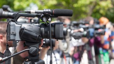 Close-up of a video camera on a tripod amidst a row of cameras, capturing an outdoor event. The setting is bright and lively with a green backdrop.