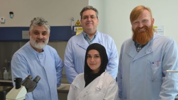 Four scientists in lab coats smile in a laboratory. A microscope is visible in the foreground, suggesting a collaborative and positive work environment.