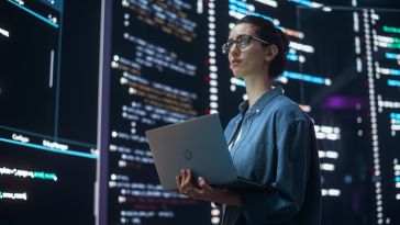 A woman engineer holds a laptop while surrounded by giant screens displaying lines of code.