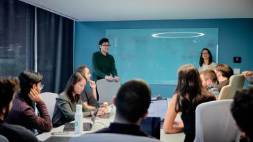 Two Kensho team members stand by a whiteboard during a team discussion while several colleagues gather at a table