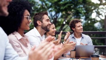 A diverse group of smiling colleagues sit at an outdoor table and applaud during a meeting or presentation.