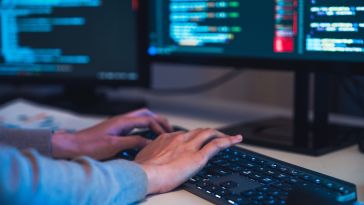 A person's hands are shown typing on a backlit keyboard in a dimly lit room, with lines of colorful computer code visible on dual monitors in the background.
