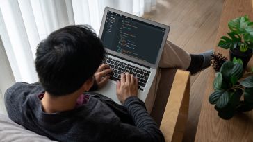 An over-the-shoulder view of a person sitting in a wooden lounge chair and typing code on a laptop in a bright, modern room with indoor plants.
