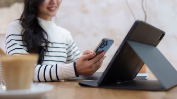 A woman in a striped sweater sits at a wooden table in a cafe, smiling while looking at her smartphone next to an open tablet and a cup of coffee.