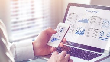 A close up of a person holding a smartphone over a laptop with various charts and graphs on both device screens.