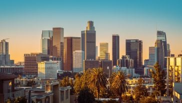 A view of the Los Angeles skyline at sunset featuring several tall skyscrapers and palm trees.