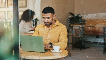 A man wearing a yellow shirt and wired earbuds works intently on a laptop at a café table with a cup of coffee.