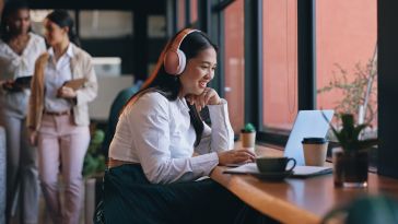 A smiling woman wearing pink headphones sits at a wooden counter by a window, working on her laptop with coffee nearby.