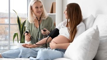 A smiling nurse measures a pregnant woman's blood pressure in a bright, cozy bedroom. The mood is warm and caring, with natural light streaming in.