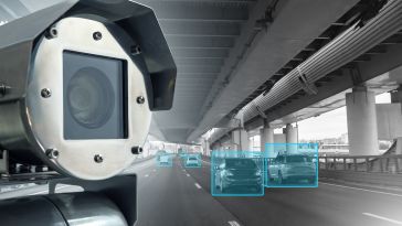 Traffic surveillance camera monitoring cars under a highway overpass. Vehicles are highlighted with blue rectangles, suggesting digital tracking.