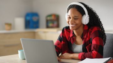 A woman in a red flannel shirt and white headphones smiles while using a laptop at a kitchen table. A notebook and mug are nearby, creating a cozy study atmosphere.
