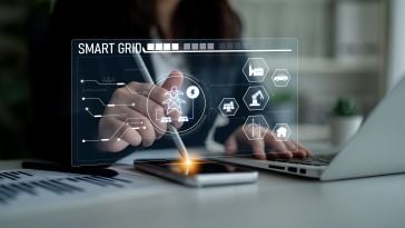 A person interacts with a digital smart grid interface on a smartphone, with icons representing energy and technology, next to a laptop on a desk.