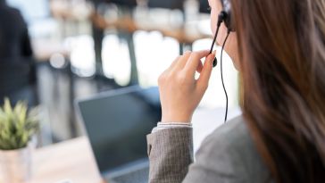 Woman in a gray blazer wearing a headset, facing a laptop in an office setting. She appears focused, suggesting a call center or customer service role.