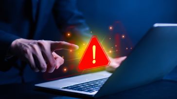 A person in a suit points at a laptop, with a red warning triangle and exclamation mark glowing above the screen, indicating a cybersecurity alert.