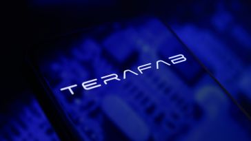 The Terafab logo is displayed on a smartphone screen placed on a reflective surface with blue light projections.