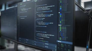 A close-up, angled view of a computer monitor displaying lines of code in a dark-themed integrated development environment (IDE) within a modern office setting.