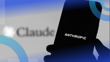 A hand holds a smartphone with the Anthropic logo in front of a screen advertising the company's Claude tool.