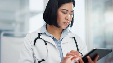 A female doctor with a white coat and stethoscope looks down intently while interacting with a digital tablet in a bright clinic setting.