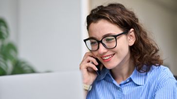 A smiling woman talking on a cellphone and looking at a computer screen