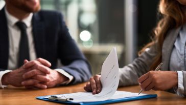 A close-up shot of two people in business attire at a wooden table, where one person is signing a document on a blue clipboard while the other sits across with hands clasped.