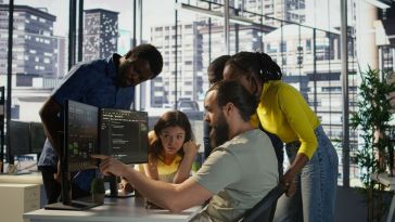 Five software engineers view data on a computer monitor while working on an AI project together in an office