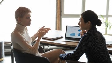 Two employees sit at a desk engaging in a serious conversation.