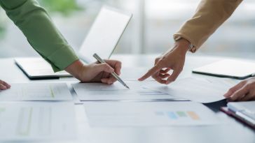 Two people collaborate around a desk, pointing to and reviewing financial charts and documents with a pen.