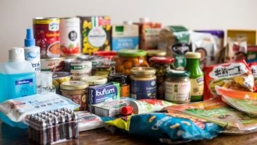 Consumer packaged goods laid out on table. Includes hand sanitizer, pills, ramen, canned goods and more.