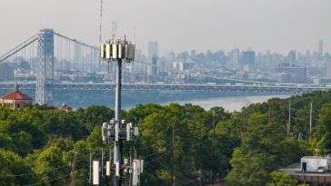 Cell tower in the foreground overlooking a tree-lined road, with the George Washington Bridge and the Manhattan skyline in the hazy background.