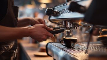 A close-up shot shows a barista's hands attaching a portafilter to a professional espresso machine as steam rises from a dark cup of coffee sitting on the drip tray.