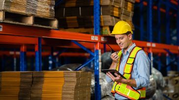 A smiling warehouse worker wearing a yellow hard hat and a high-visibility safety vest looks at a tablet while speaking into a walkie-talkie amid rows of inventory shelving.