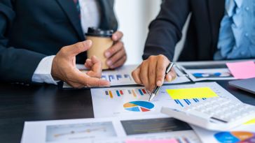 Two business professionals in suits review financial data together at a desk covered in charts, graphs, sticky notes, and a calculator, with one person pointing a pen at a pie chart while the other gestures during the discussion.