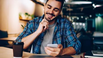 Man smiling while looking at a smartphone in a cafe, with a coffee cup and laptop on the table.