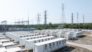 An aerial view of a battery storage facility featuring rows of white containerized battery units situated next to an electrical substation and high-voltage transmission towers.