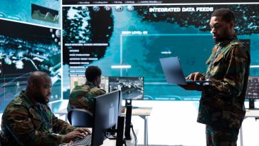  Three military personnel in camouflage uniforms monitor high-tech digital displays and work on laptops in a sophisticated, blue-lit command center featuring integrated data feeds and satellite imagery.