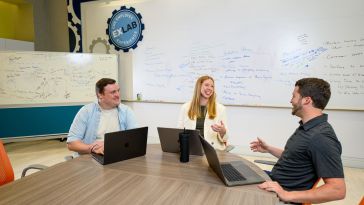 Three Cox Automotive employees sit at a table with their laptops in front of a whiteboard