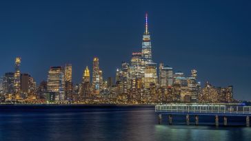Lower Manhattan skyline at night, with One World Trade Center illuminated above the Hudson River and a pier in the foreground.